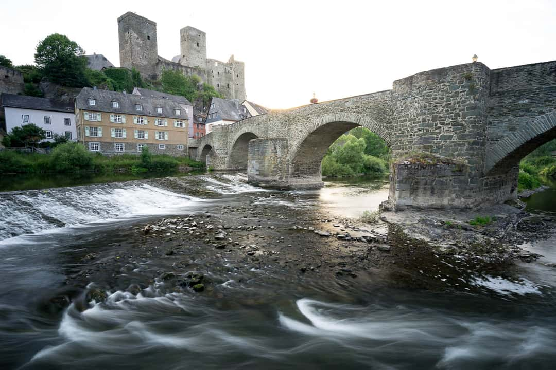 With polarizer: Instead of having a bright foreground, we now see nice little waves, that lead the eye of the viewer from the bright castle in the background through the foreground towards the bridge on the right side. The stones in the riverbed are some nice additional details, that are visible now.